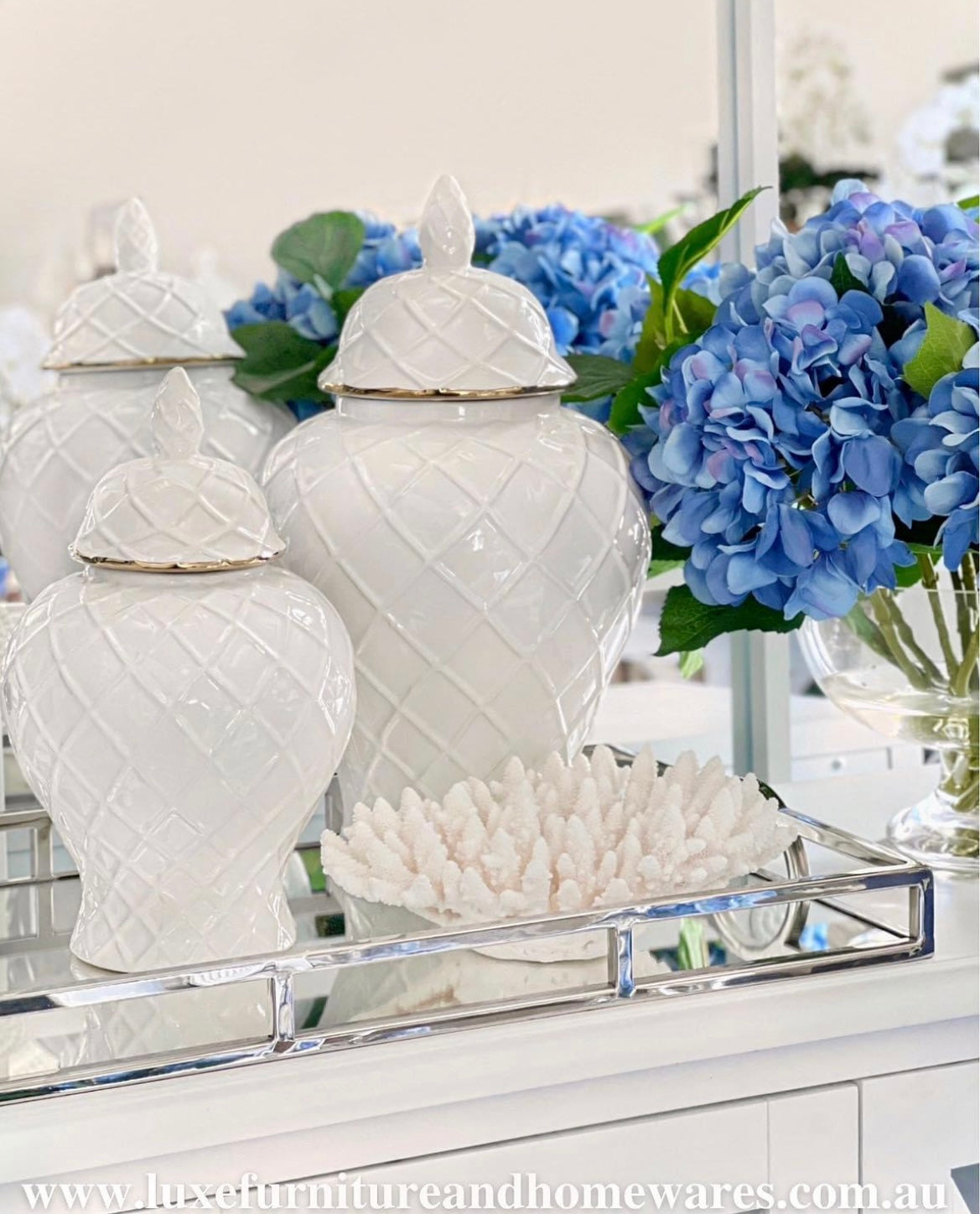A white ceramic ginger jar with a gold trimmed lid, featuring a criss-cross raised diamond design, placed on a surface with other decorative items and a blue floral arrangement in the background.