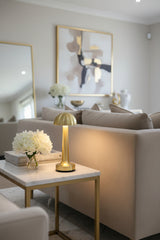A gold USB touch lamp with a round top, placed on a reflective surface with a white flower arrangement in the background.